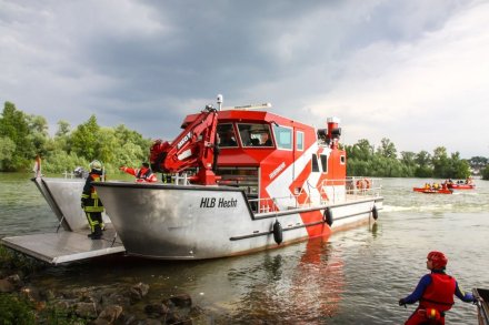 Rotes Feuerwehrboot mit Namen Hecht, ein Hilfeleistungslöschboot (HLB), mit geöffneter Laderampe auf dem Rhein macht am Land fest. Im Hintergrund sind weitere kleine Boote der DLRG zu sehen.