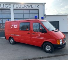 Rotes Feuerwehrfahrzeug, ein Mannschaftstransportwagen (MTW), steht vor einem Feuerwehrgerätehaus.