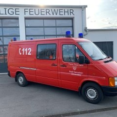 Rotes Feuerwehrfahrzeug, ein Mannschaftstransportwagen (MTW), steht vor einem Feuerwehrgerätehaus.