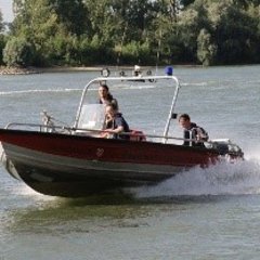 Rotes Feuerwehrboot, ein Mehrzweckboot (MZB), mit dreiFeuerwehrleuten. Das Boot fährt schnell auf dem Rhein. Gischt spritzt auf.