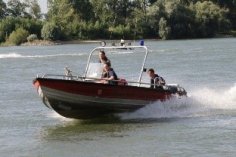 Rotes Feuerwehrboot, ein Mehrzweckboot (MZB), mit dreiFeuerwehrleuten. Das Boot fährt schnell auf dem Rhein. Gischt spritzt auf.