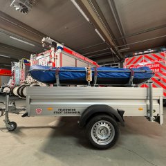 Ein Rettungsboot der Feuerwehr (RTB-1) auf einem silbernen Anhänger. Im Hintergrund die Fahrzeughalle mit verschiedenen Feuerwehrfahrzeugen.