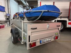 Ein Rettungsboot der Feuerwehr (RTB-1) auf einem silbernen Anhänger. Im Hintergrund die Fahrzeughalle mit verschiedenen Feuerwehrfahrzeugen.