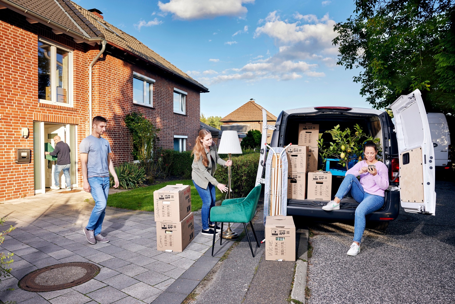 Links im Bild trägt ein Mann einen Stuhl in ein Haus. In der Bildmitte ein Mann und eine Frau die eine Lampe und Umzugskisten in der Hand halten. Rechts sitzt eine Frau mit einem Handy in der Hand in der geöffneten Hecktür eines Kleinbusses voller Umzugskisten.