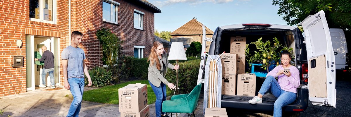 Links im Bild trägt ein Mann einen Stuhl in ein Haus. In der Bildmitte ein Mann und eine Frau die eine Lampe und Umzugskisten in der Hand halten. Rechts sitzt eine Frau mit einem Handy in der Hand in der geöffneten Hecktür eines Kleinbusses voller Umzugskisten.