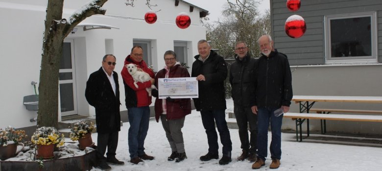 Sechs Personen stehen vor dem Gebäude des Tierschutzvereins im verschneiten Hof unter einem winterlichen Baum, der mit großen roten Kugeln geschmückt ist. Frau Martina Grundmann und Herr Bürgermeister Burger halten einen symbolischen Scheck in den Händen. Herr Jirele (zweite Person von links) hält einen weißen Hund im Arm.